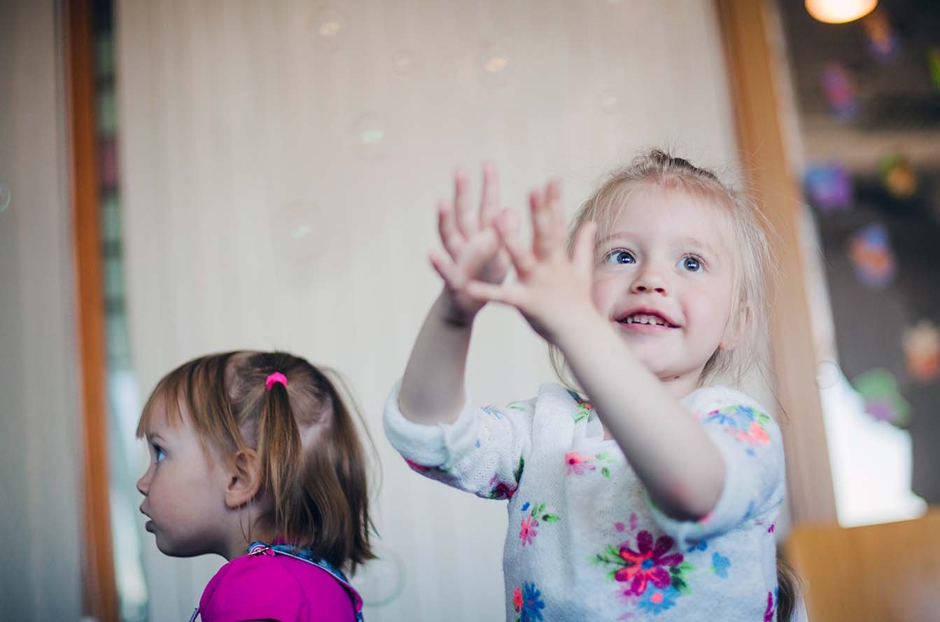Young child smiles and raises both hands while another child stands nearby.