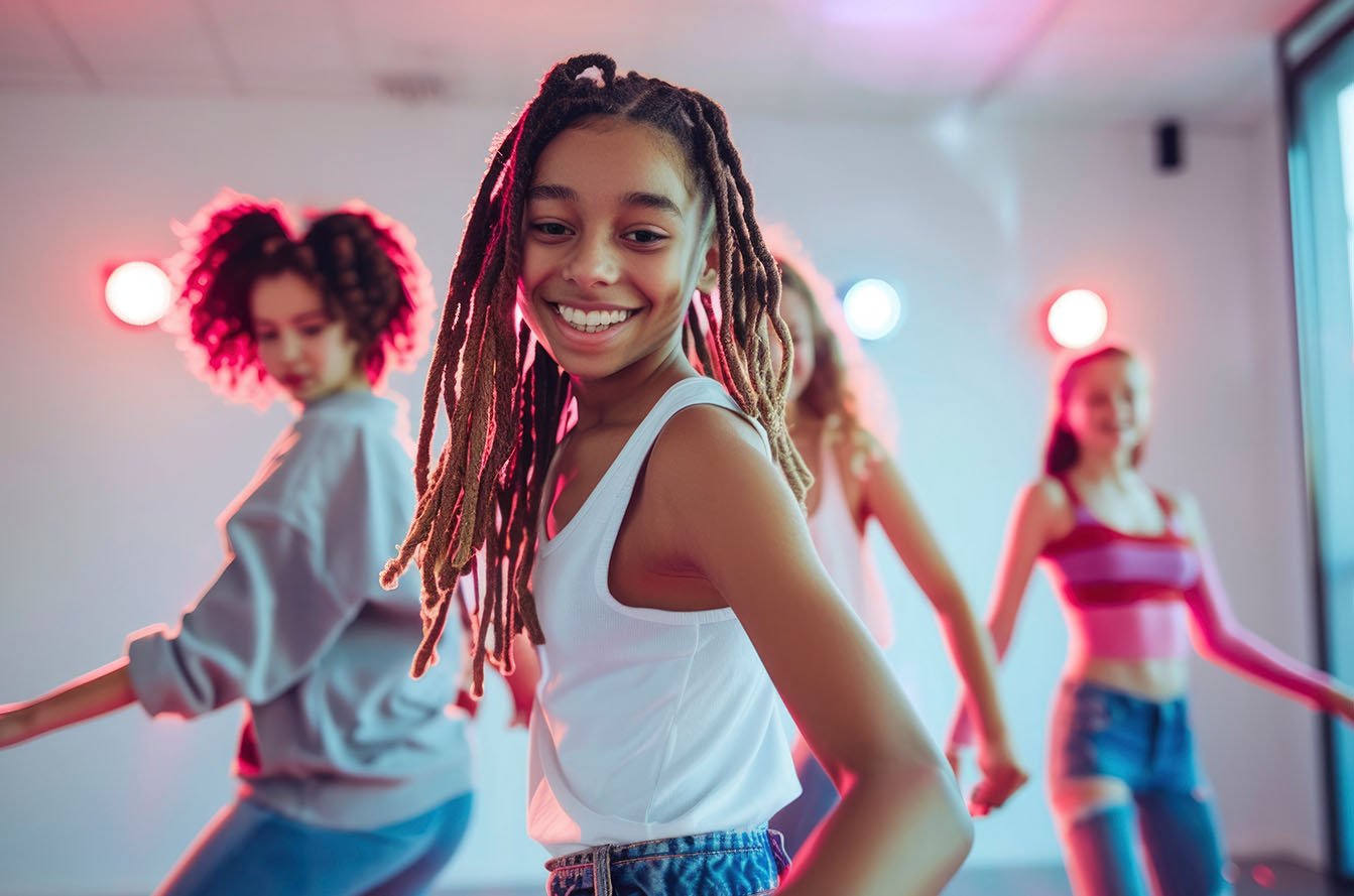 Hip Hop Classes for Kids - Confidence Through Movement Smiling young dancer with long locs dancing with a group in a studio lit with pink and blue lights.